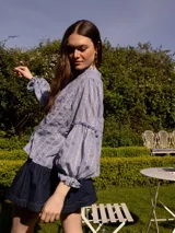 Camisa verde con rayas verticales blancas y bordados de flores. Tiene cuello mao, mangas largas abullonadas con puños elásticos y cierre frontal con botones de nácar.