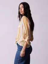 Blusa de gasa estampada a rayas verticales en tonos celeste, lila, naranja y marrón. Tiene cuello a la base con escote en V y botones. Delantera con bolsillo aplicado. Largo de manga regulable con presilla y botón. Terminación anudada opcional.