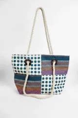 Bolso de playa o tote bag con diseño de patchwork en tonos azules, morados, naranjas y beige. Presenta secciones con lunares azules y secciones con rayas horizontales. Las asas son de cuerda trenzada color crema.