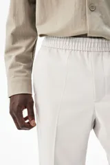 Pantalón de vestir gris claro, de corte slim fit, con cintura elástica y cordón ajustable. Presenta bolsillos laterales y traseros ribeteados, pliegues cosidos y doblez en el bajo.