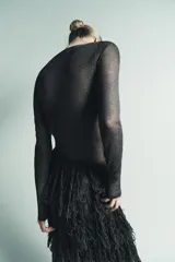 Vestido midi negro, semitransparente, de manga larga y cuello redondo. La parte superior presenta un tejido de punto fino con costuras que simulan un bustier. La falda está compuesta por capas de flecos largos y densos.