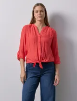 Blusa color salmón de gasa marca Zac & Rachel, con cuello a la base, escote en V y abertura central con botones. Tiene un bolsillo aplicado en la delantera y mangas 3/4 regulables con presilla y botón. El ruedo puede atarse con un nudo.