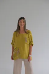 Blusa color mostaza de corte holgado, con cuello redondo, mangas 3/4 con botón para sujetar y detalle de bolsillo y aplique floral en la parte inferior.