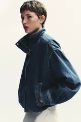 Chaqueta corta de denim azul con cuello alto, manga larga con hombros caídos, bolsillos laterales, bajo con elástico y cierre frontal con cremallera y botones ocultos.