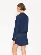 Chaqueta de denim color crudo, corte a la cintura y mangas largas con volados en puños y ruedo. Cierre frontal con broches dorados y bolsillos con broches dorados.
