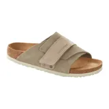 Sandalia de cuero gamuzado en color taupe, modelo Kyoto de Birkenstock. Presenta un diseño de una sola tira ancha envolvente con cierre de velcro ajustable y remaches metálicos decorativos. Cuenta con la clásica plantilla anatómica de corcho y látex, y suela de EVA en color blanco.