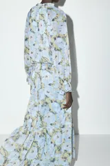 Vestido largo con estampado floral en tonos celeste, amarillo y marrón. Tiene cuello redondo con volantes y lazada, escote pico, manga larga con puño elástico con volantes, forro interior y cierre frontal con botones.