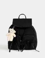 Mochila negra de tela deportiva con cierre de cordón ajustable y solapa con broche a presión. Tiene manija superior, correas regulables, doble bolsillo frontal con cierre y adorno de oso de peluche colgando.