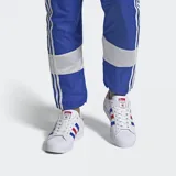 Championes Adidas Superstar blancos con detalles en azul y rojo. Presentan las tres tiras características de Adidas en los laterales en azul y rojo, y refuerzo de talón en rojo con la palabra "STAR" en blanco. La puntera es de goma con textura.