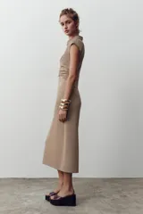Vestido midi color beige con cuello polo, escote en pico, manga sisa caída y detalle de drapeado lateral. Cierre frontal con botones metálicos en relieve.
