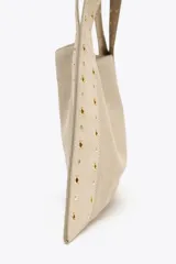 Bolso de hombro color beige, confeccionado en serraje, con tachas metálicas doradas en el cuerpo y las asas. Cuenta con un bolsillo interior y cierre mediante botón imantado.