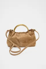 Bolso mini tipo saca de cuero serraje color marrón con efecto soft. Tiene doble asa de mano metálica dorada y asa bandolera extraíble. Cuenta con bolsillo interior con cremallera y cierre mediante fruncido y botón imantado.