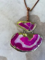 Collar con colgante de dos piedras naturales irregulares engarzadas en metal plateado y dorado, suspendidas de un cordón de cuero marrón con detalle de nudo corredizo. Las piedras tienen vetas en tonos naranja, marrón y blanco.