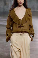 Blusa color marrón con escote en pico, manga larga abullonada con elástico en los puños y volantes en el escote y mangas.