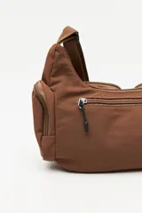 Cartera tipo shoulder color beige, de diseño utilitario y formato curvo. Confeccionada en material liviano y resistente, cuenta con correa regulable para llevar al hombro o cruzada. Presenta múltiples bolsillos con cierre en el frente y laterales, y un parche con el logo "Piece & Cake" en el frente.