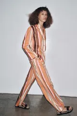 Pantalón de tiro medio con estampado de rayas verticales en tonos marrón, naranja, rosa y beige. Cuenta con trabillas en la cintura, cinturón ajustable con lazadas, bolsillos laterales y cierre frontal con cremallera y botones.