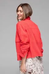 Campera corta color coral con cuello camisero, cierre frontal con botones, bolsillos de parche en el pecho y mangas largas con puños abotonados. Presenta calados tipo broderie en hombros, mangas y espalda.