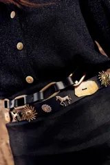 Cinturón negro de cuero con apliques de tachas plateadas y doradas con forma de soles, caballos y flores. Se ajusta con un lazo en la parte posterior.