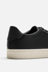 Zapatilla urbana de cuero sintético color negro, con cordones y suela de goma color crema.