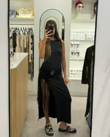 Vestido largo negro sin mangas, con corte ajustado en la parte superior y falda con abertura lateral alta y detalle de flecos. Incluye cinturón negro con hebillas plateadas.