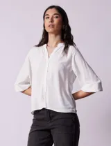 Blusa blanca de gasa marca Sioni, con cuello a la base y escote en V. Tiene abertura central con botones y mangas 3/4 con terminación fruncida.