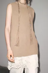 Vestido color beige, de corte recto, con cuello redondo y sin mangas. Presenta un tejido de punto en la parte superior y un volante de encaje blanco en el ruedo.