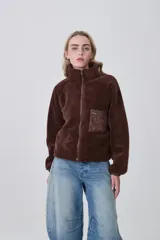 Campera de poliéster tipo sherpa en color natural, con diseño holgado, cuello alto y cierre frontal completo. Presenta un bolsillo de parche con cierre en el pecho y mangas largas con puños elásticos.