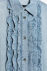 Camisa de denim celeste con cuello solapa y manga corta. Presenta detalle de volantes en el frente y bajo asimétrico con aberturas laterales.