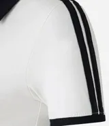 Blusa polo de malla de algodón color blanco con cuello y mangas cortas con ribete negro. Presenta bordado de moña en el pecho y dos rayas negras en cada manga.