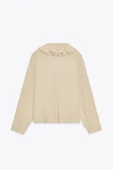 Camisa color beige de algodón con escote en V y volantes. Tiene mangas largas con botón en los puños y cierre con lazos en el frente.