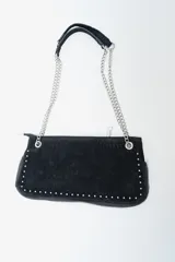 Cartera de mano tipo clutch o baguette, color negro, con textura de gamuza o terciopelo. Presenta tachas metálicas plateadas a lo largo de los bordes inferiores y laterales. Tiene una correa corta de hombro compuesta por una tira de cuero sintético negro y cadenas metálicas plateadas.