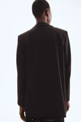 Blazer largo negro, de corte recto y botonadura sencilla con dos botones frontales. Presenta solapas de muesca, bolsillos delanteros ribeteados con solapa y hombreras.