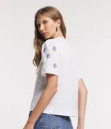 Remera blanca de algodón con cuello redondo, mangas cortas abullonadas y pequeños bordados florales azules en las mangas.