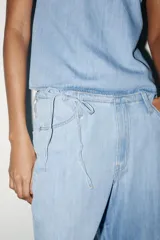 Pantalón denim celeste de tiro medio con cintura ajustable con cordones, bolsillos laterales y de plastrón en espalda. Cierre frontal con cremallera y gancho metálico.