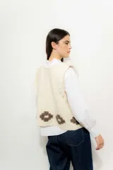 Chaleco tejido color crema con cuello redondo y borde con flecos. Presenta apliques de flores marrones en la parte inferior.