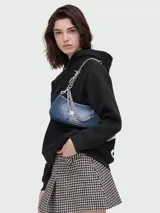 Bolso de hombro Marc Jacobs modelo The Shoulder Bag Charm, confeccionado en denim cepillado azul. Presenta una correa de cadena plateada con dijes colgantes, incluyendo una estrella y un alfiler de gancho. El bolso tiene forma curva y un cierre superior.