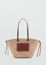 Bolso tipo shopper de paja trenzada color beige con aplique rectangular de cuero marrón con tachas doradas. Tiene doble asa de cuero marrón y cordones laterales ajustables.