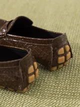 Mocasín de cuero vacuno color marrón con textura croco y acabado brillante. Incluye aplique metálico en forma de corazón sobre el frente y suela de goma.
