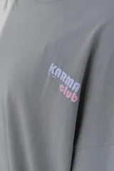 Remera gris de corte oversize con cuello redondo y estampado pequeño en el pecho con la frase "Karma Club" en color lila.