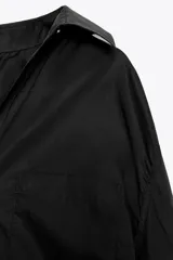 Camisa negra de cuello solapa y manga larga amplia. Tiene un bolsillo de parche en el delantero, detalle de pliegues y cierre frontal con botones.