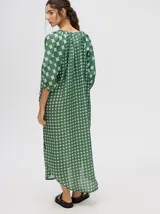 Vestido largo con mangas 3/4 y cuello mao, confeccionado en tela con estampado de círculos en tonos verde oliva y celeste pálido. Presenta cierre frontal de botones marrones y corte amplio con aberturas laterales.