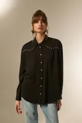 Camisa holgada de manga larga, color negro, con cuello camisa y botones frontales. Presenta apliques de tachas plateadas en los hombros y a lo largo de la tapeta frontal.