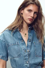Mono corto de denim azul, con cuello solapa, manga corta, bolsillos delanteros y traseros, cintura elástica en la espalda y cierre frontal con cremallera y botones metálicos.