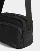 Bandolera negra de tela deportiva con bolsillo frontal con cierre, correa regulable y tag de goma.
