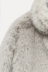 Abrigo corto de pelo sintético color gris claro, con cuello subido y manga larga. Tiene bolsillos laterales ocultos en la costura, forro interior combinado y cierre frontal con ganchos metálicos forrados.
