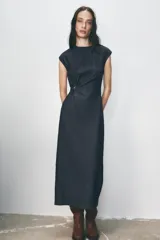 Vestido midi de denim azul oscuro, con cuello redondo y sisas. Presenta un aplique dorado en la cintura y abertura en la espalda. Cierre trasero con cremallera oculta.