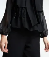 Blusa negra de chiffon con cuello alto con lazo, mangas largas con puños ajustados, detalle de volado delantero y volado en encaje.