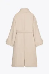 Abrigo largo color beige confeccionado en hilatura con lana. Tiene cuello subido, manga larga con trabillas, bolsillos laterales y cinturón ajustable con lazada. El cierre frontal es cruzado con botones.