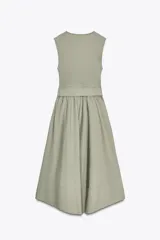 Vestido midi color verde oliva, de cuello redondo y sin mangas. Presenta un cinturón con hebilla metálica y detalle de volantes en la falda.