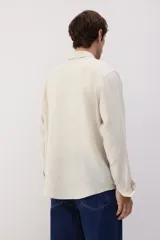 Camisa de lino color crema, de corte recto, con cuello inglés, tapeta clásica, mangas largas con puños abotonados y bajo redondeado.